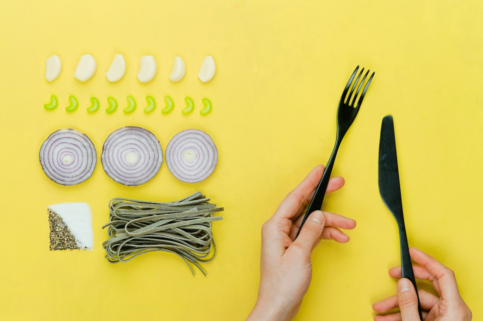 Neatly arranged ingredients on yellow background include garlic, celery, onion, spices, and pasta. Hands hold black fork and knife, evoking minimalist style.