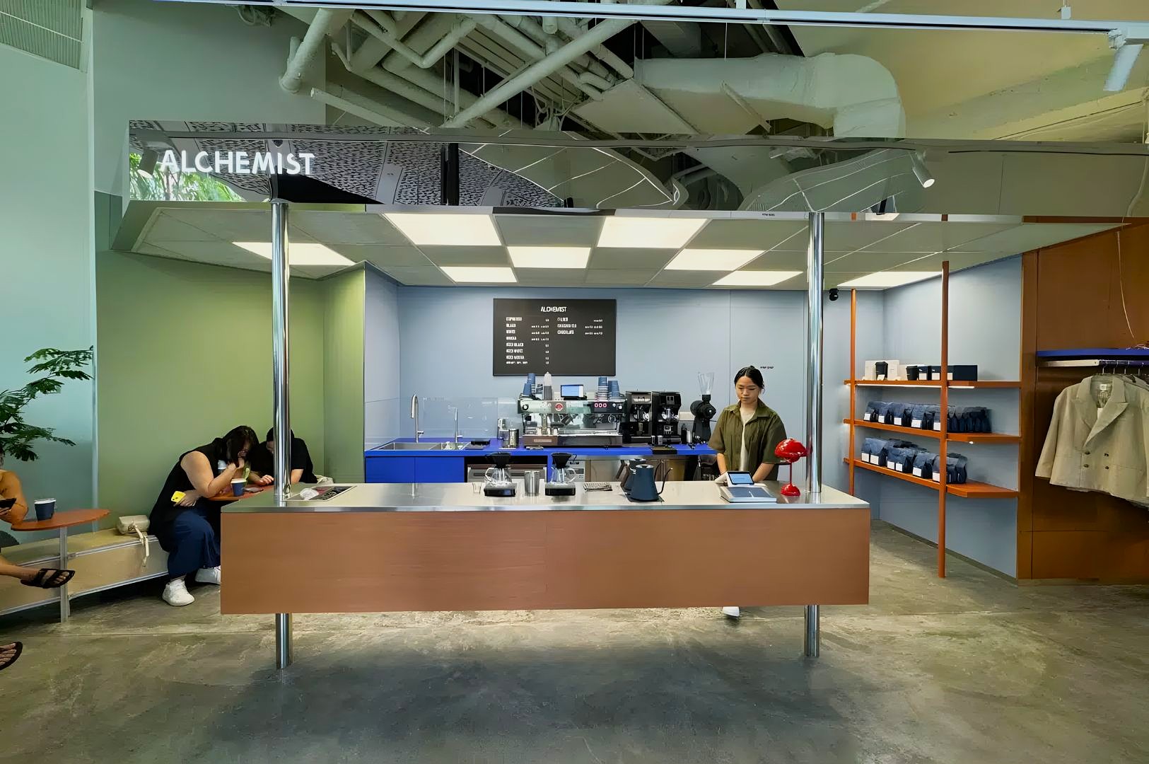 Modern cafe "Alchemist" with a minimalist design. A barista stands behind a counter, surrounded by coffee equipment. Shelves display goods.
