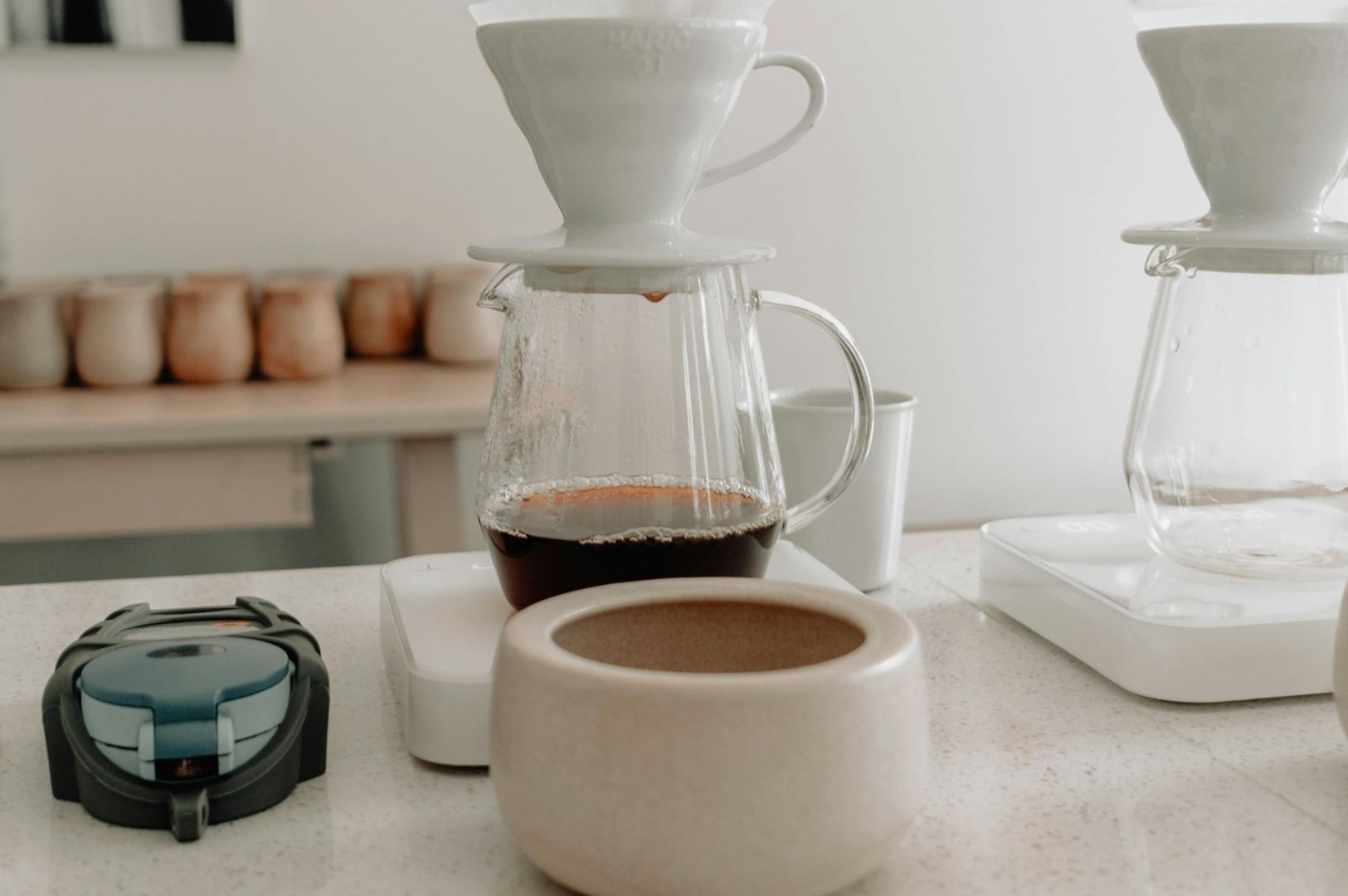 Pour-over coffee setup on a white counter with glass carafes, ceramic coffee drippers, a simple scale, and assorted mugs in the background. Cozy ambiance.