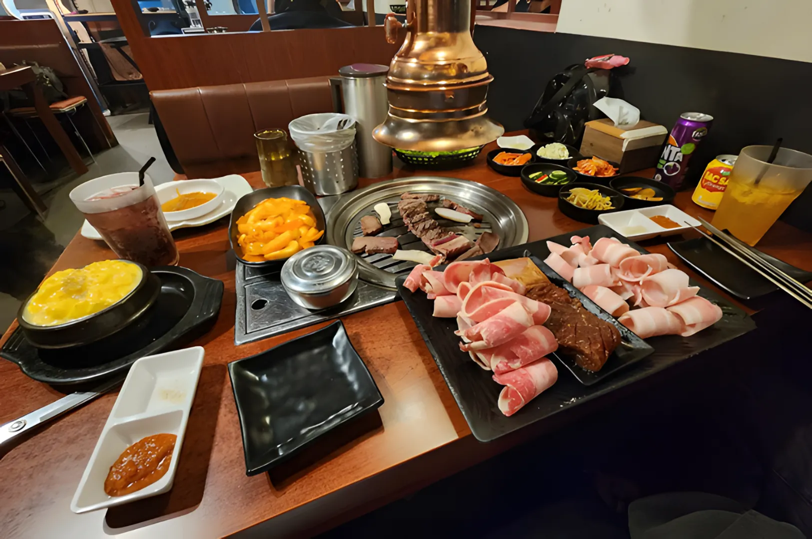 A Korean barbecue setup with raw meats ready to grill, assorted side dishes, dipping sauces, and drinks on a wooden table. A warm, cozy atmosphere.
