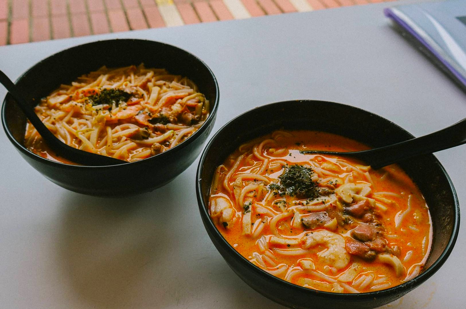 Two black bowls of creamy ramen soup with noodles, shrimp, mushrooms, and greens are on a light table. A warm, inviting meal scene.