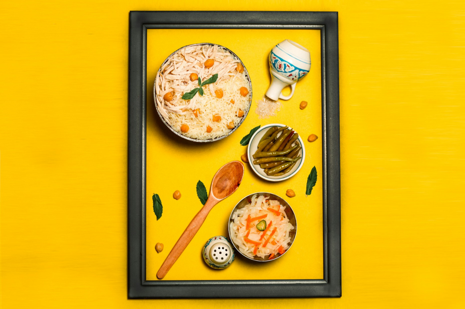 A framed arrangement of a rice dish, pickled vegetables, and green beans on a bright yellow surface. Wooden spoon and small jar add a rustic touch.