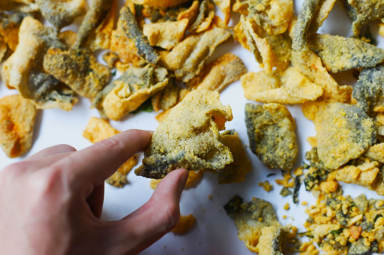 A close-up of a hand lifting a crumb-coated, fried snack piece from a scattered tray, the scene highlighting salted egg fish skin crisps among other pieces.