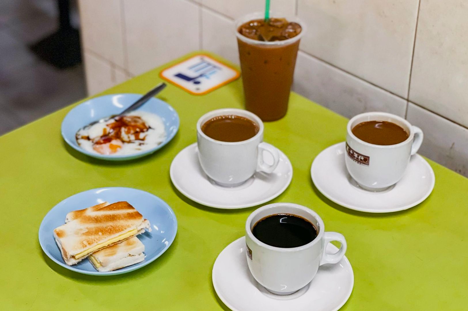 A table with three cups of coffee, iced coffee, soft-boiled eggs in a bowl, and kaya toast on a pastel green surface, creating a warm, inviting atmosphere.