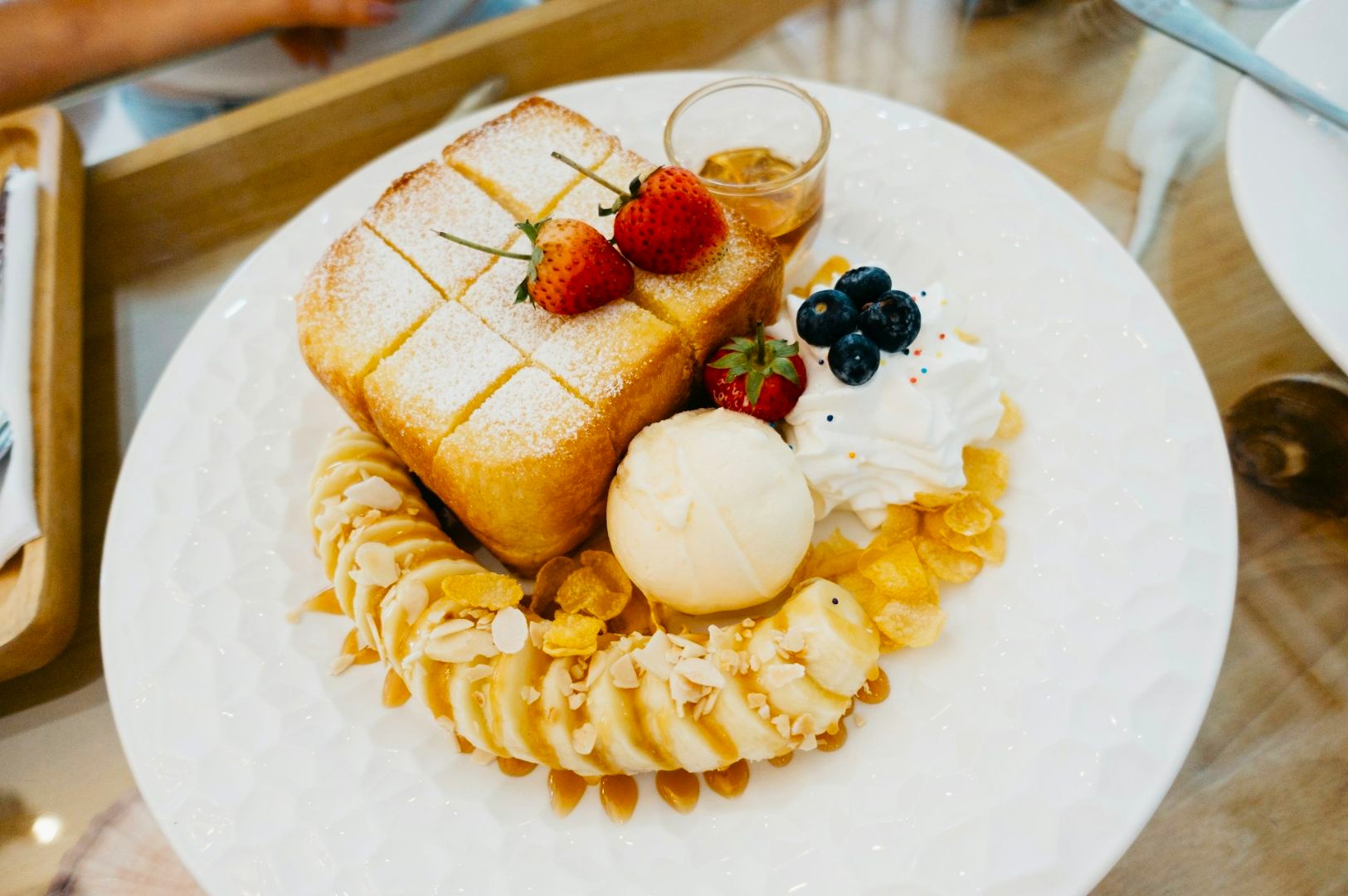 A plate with honey toast topped with strawberries and powdered sugar, surrounded by banana slices, almond flakes, whipped cream, blueberries, and ice cream.