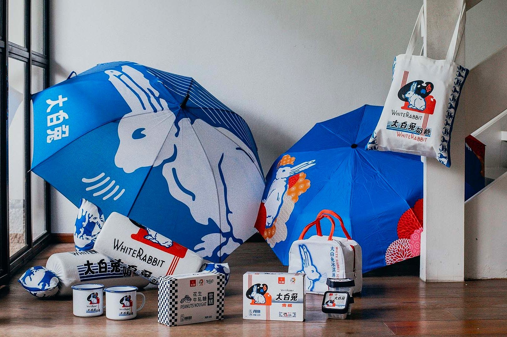 Two blue umbrellas featuring a white rabbit design are surrounded by White Rabbit branded items, including a tote bag, mugs, and boxes, set on a wooden floor.