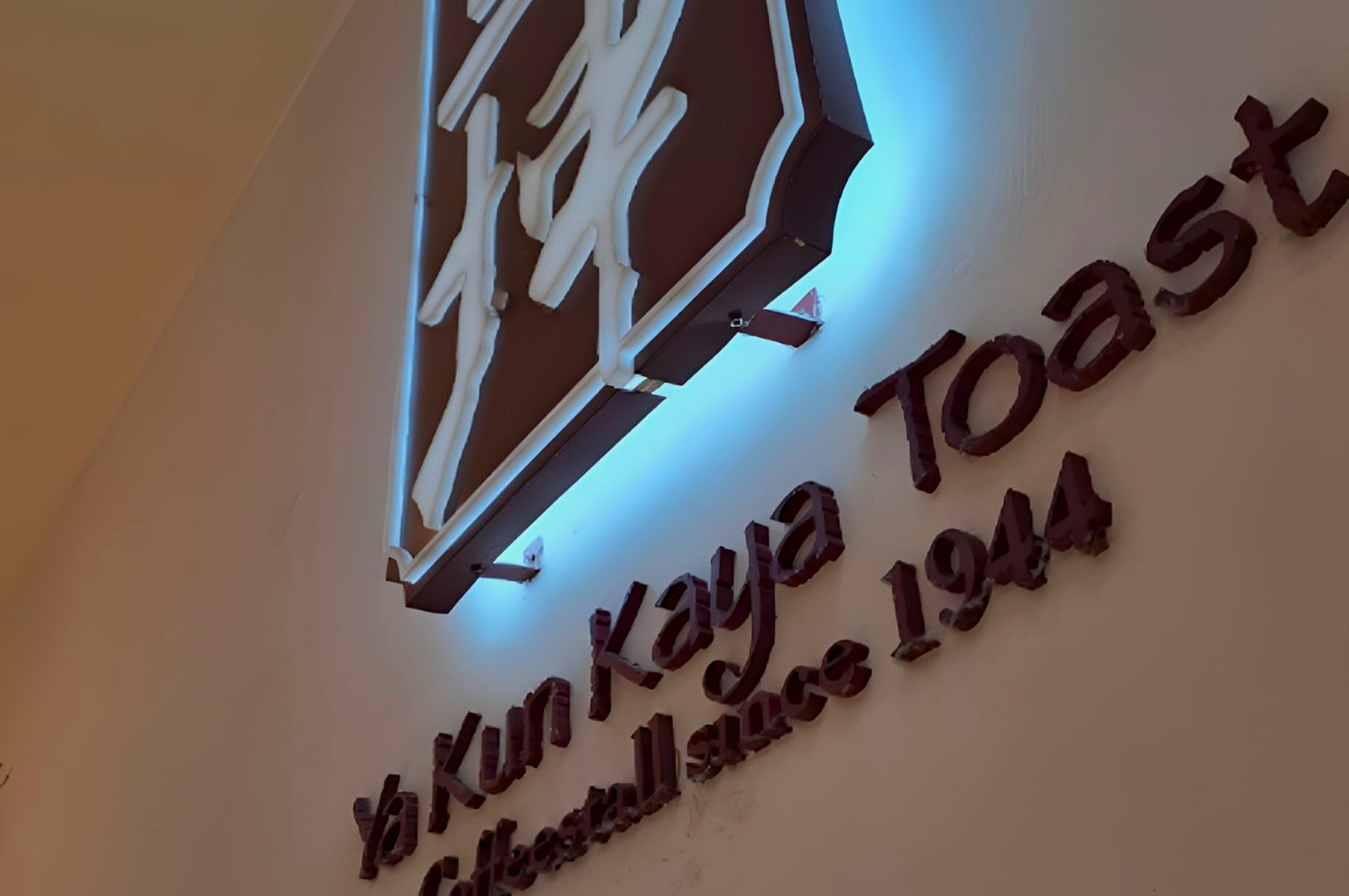 Wall signage in a cafe featuring illuminated Chinese characters above the words "Ya Kun Kaya Toast, Established since 1944," conveying tradition.