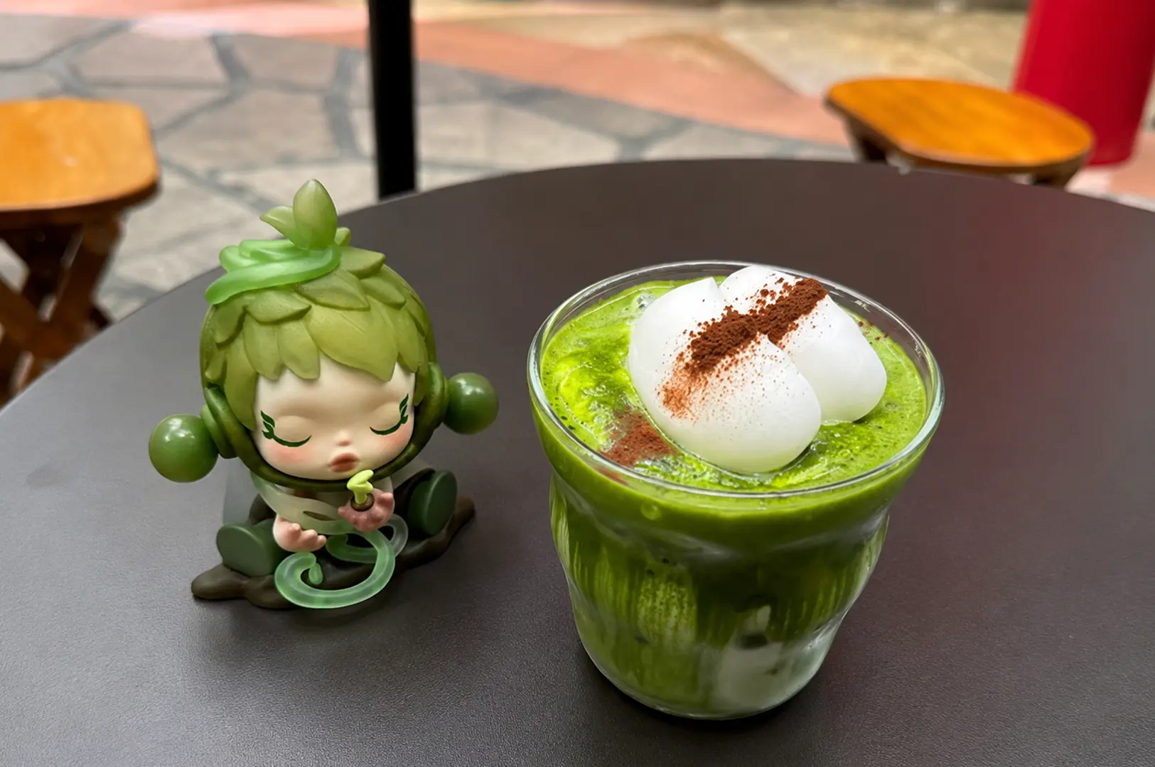 An iced matcha latte topped with white mochi cream and a dusting of cocoa powder, served on a cafe table next to a green Dimoo designer toy figure.