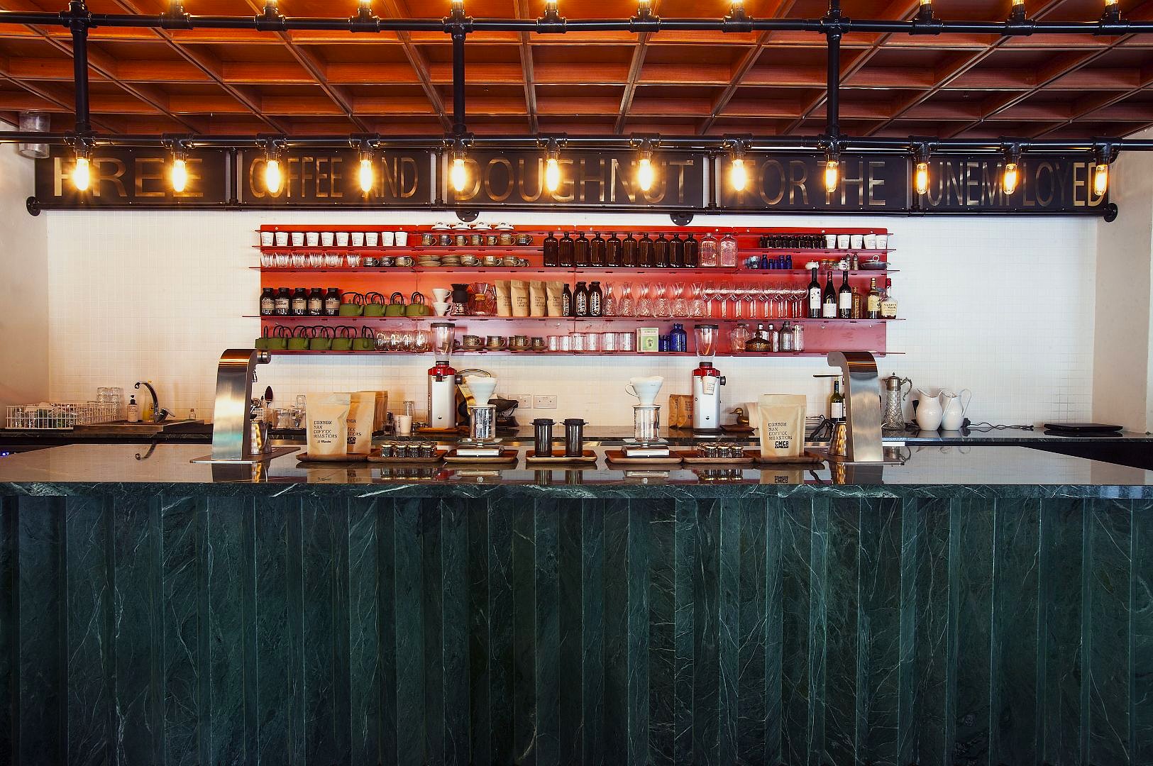 Green marble countertop bar with a vintage vibe. Shelves behind hold coffee supplies and bottles. Overhead sign reads, “Free coffee and doughnuts for the unemployed.”