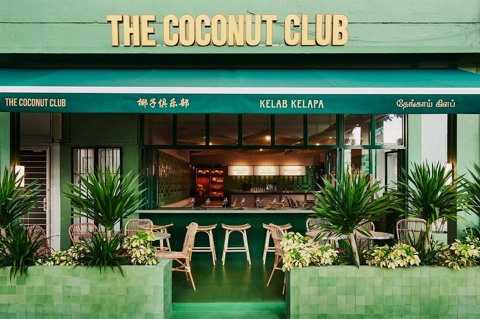 Charming café exterior with green tones, named "The Coconut Club." Features fresh plants, wicker chairs, and a cozy, inviting atmosphere.