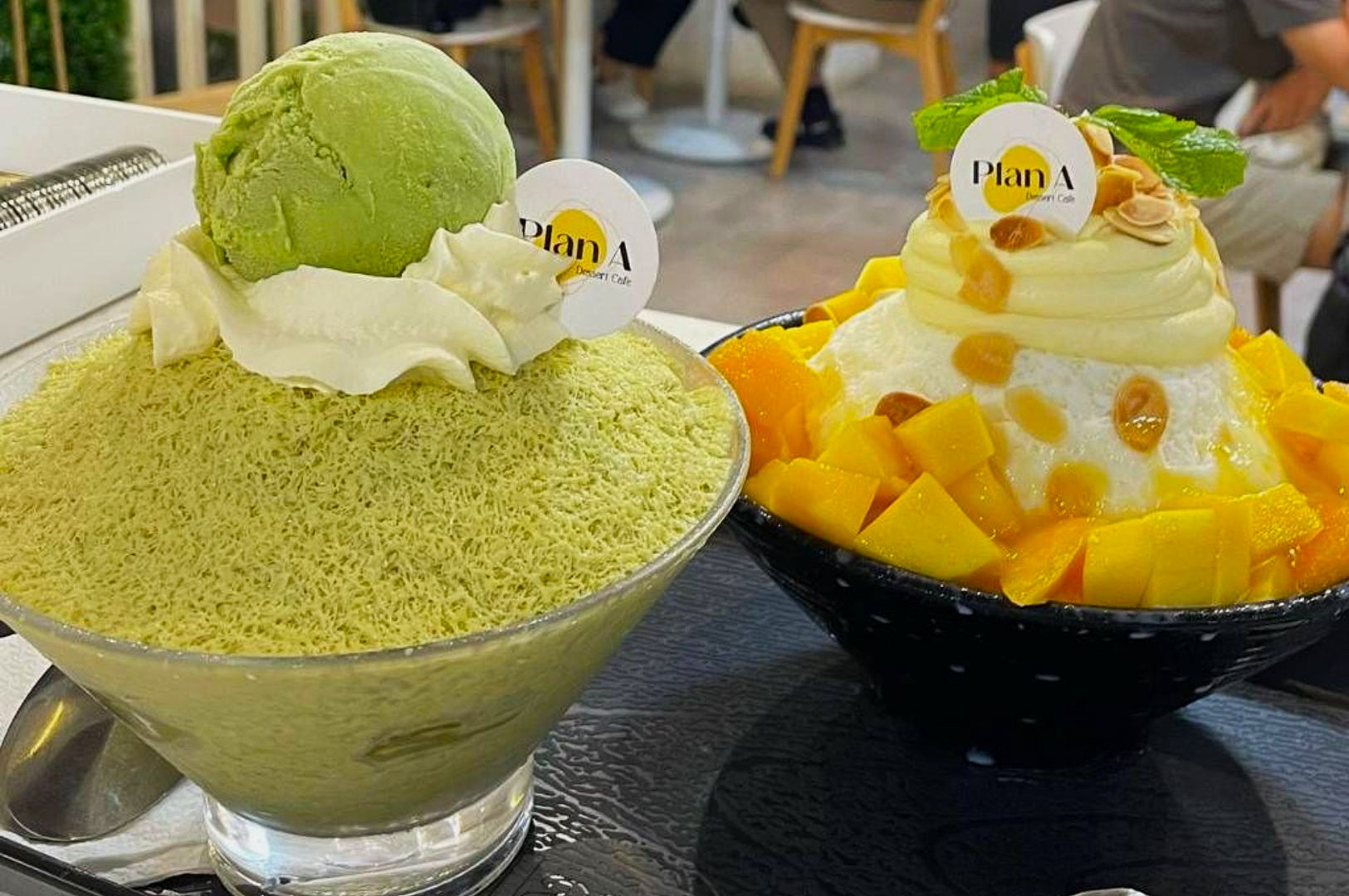 Two bowls of shaved ice desserts are on a table. The left bowl features green tea shaved ice with green ice cream. The right bowl has milk shaved ice topped with mango chunks and nuts. Both are garnished with whipped cream and mint leaves, creating a colorful and refreshing presentation.
