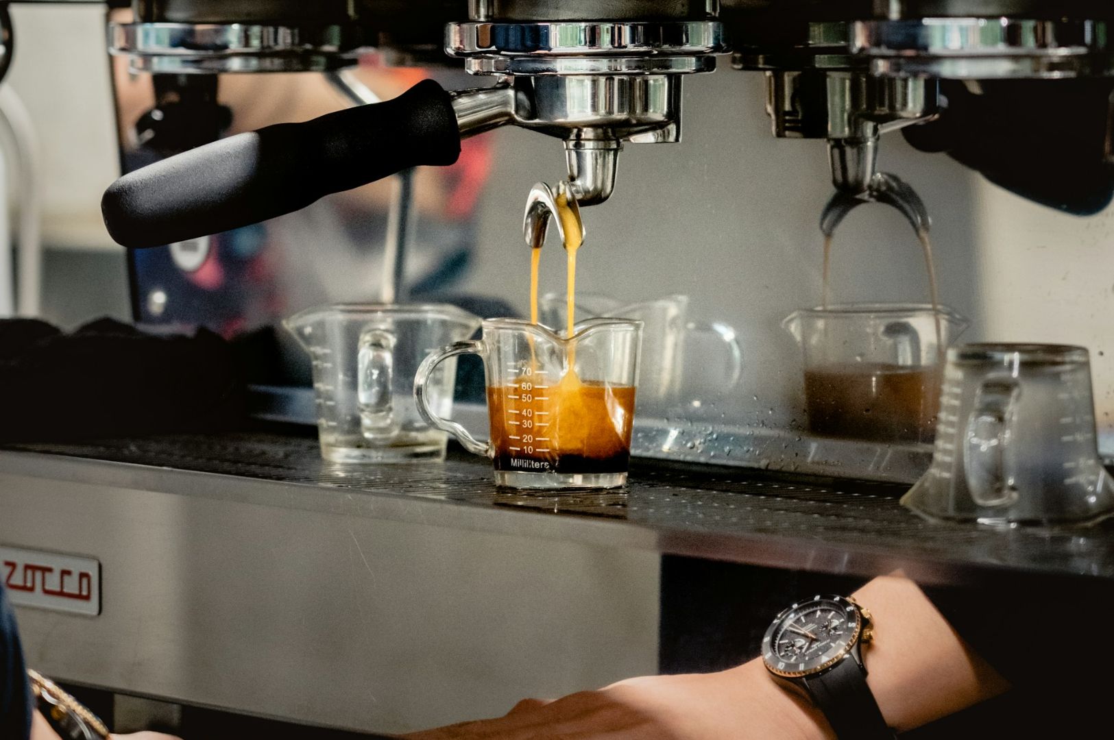 Espresso machine pouring coffee into a glass measuring cup. A person's hand, wearing a watch, adjusts the machine. The setting is a busy café.