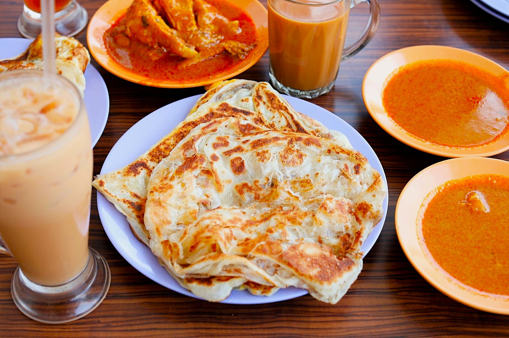 A spread of Malaysian food on a wooden table features crispy roti canai on a white plate, curry bowls, tea, and iced drinks, creating a warm, inviting scene.