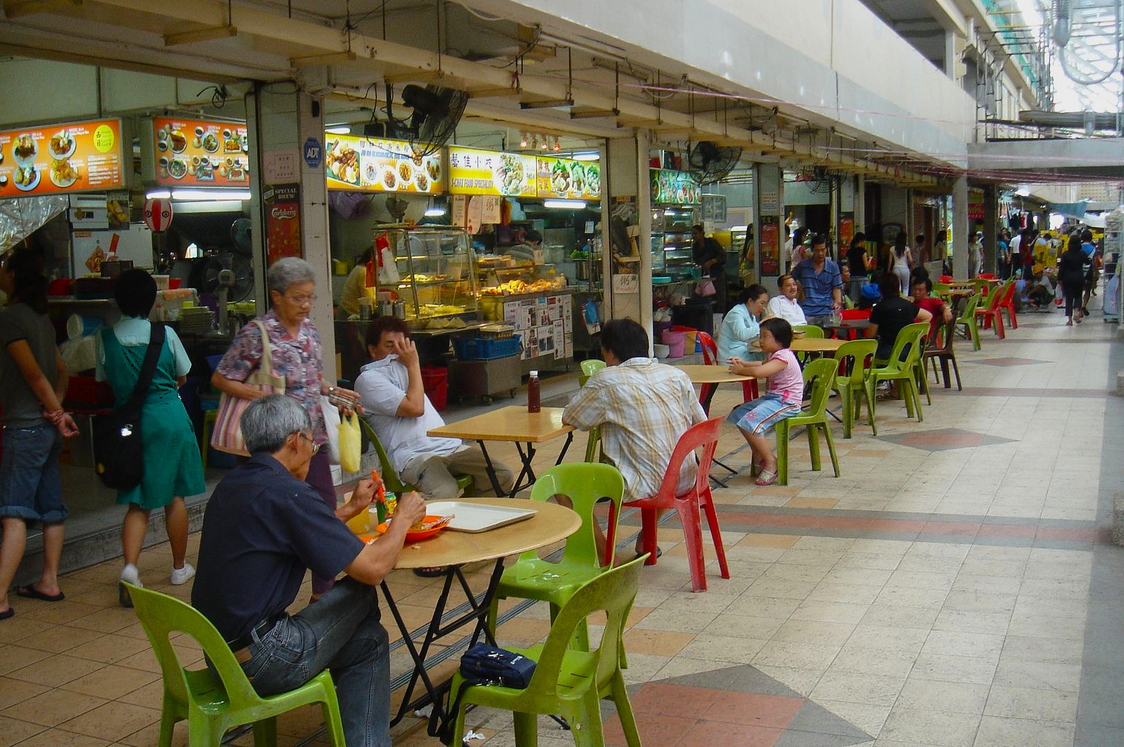 Bustling food market with colorful chairs and tables lining the corridor. People are eating and chatting. Food stalls display vibrant menus. Relaxed atmosphere.