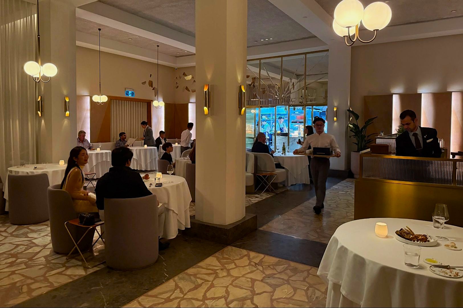 A stylish, dimly-lit restaurant with elegant decor, featuring round tables with white tablecloths, patrons dining, and attentive waitstaff serving dishes.
