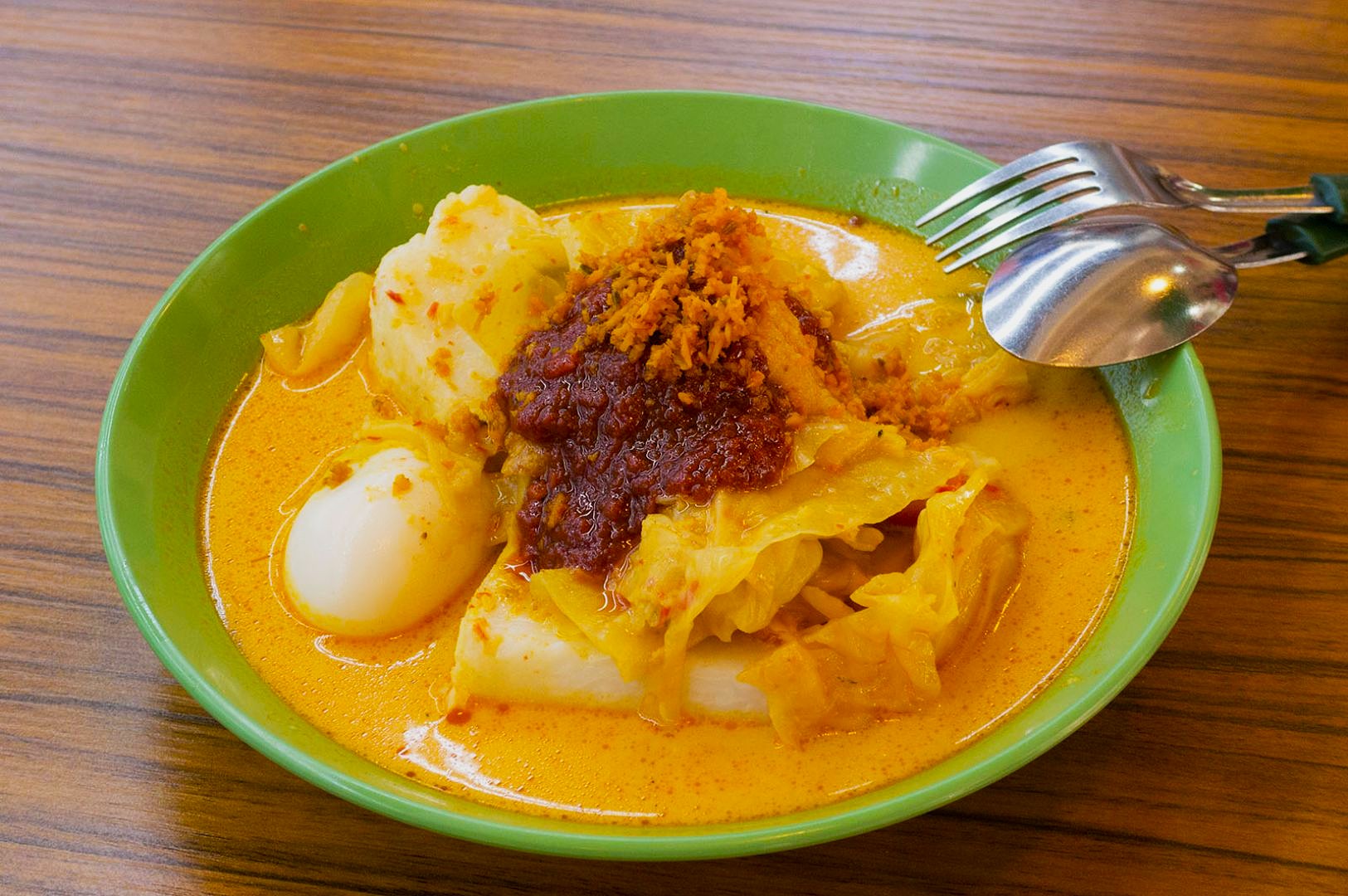 A bowl of lontong, featuring compressed rice cakes, boiled egg, cabbage, and sambal in a rich, creamy curry. Utensils rest on the bowl's edge.