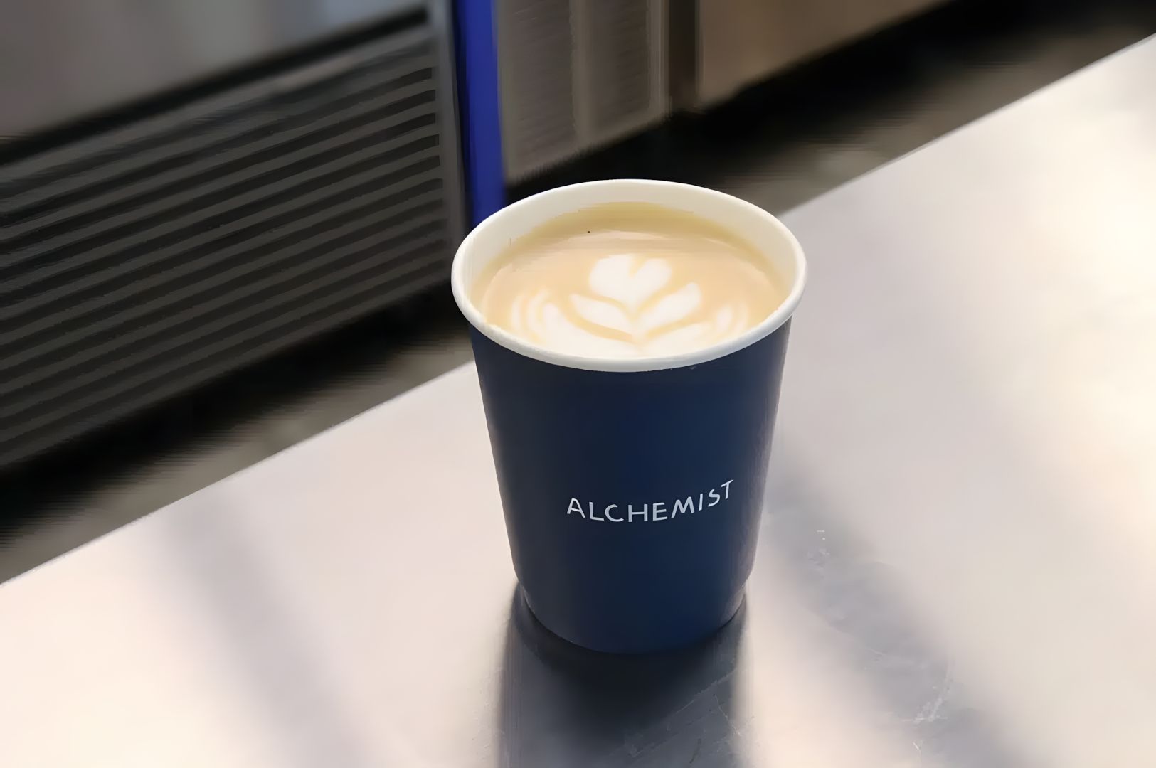 A blue paper cup labeled "Alchemist" on a metal counter holds a latte with delicate leaf-shaped latte art, conveying a cozy and inviting atmosphere.