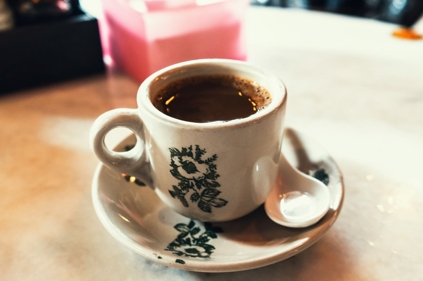 A ceramic cup of coffee sits on a saucer with a small white spoon. The cup and saucer feature green floral patterns, creating a cozy, inviting feel.
