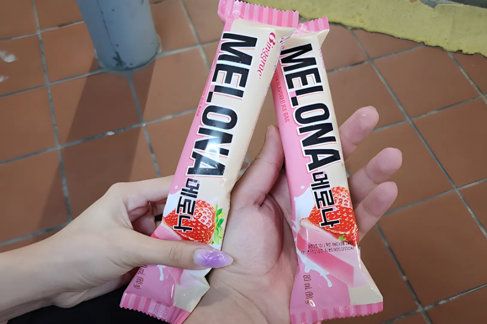 Two hands holding Melona strawberry-flavored popsicle packages. The background shows tiled flooring, creating a casual, relaxed setting.