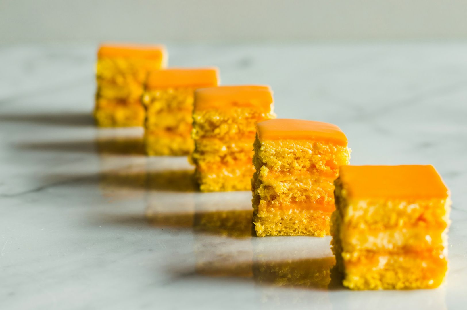 Five orange-glazed cake squares are aligned diagonally on a marble surface, each slice showcasing layered textures. The setting gives a modern, appetizing feel.