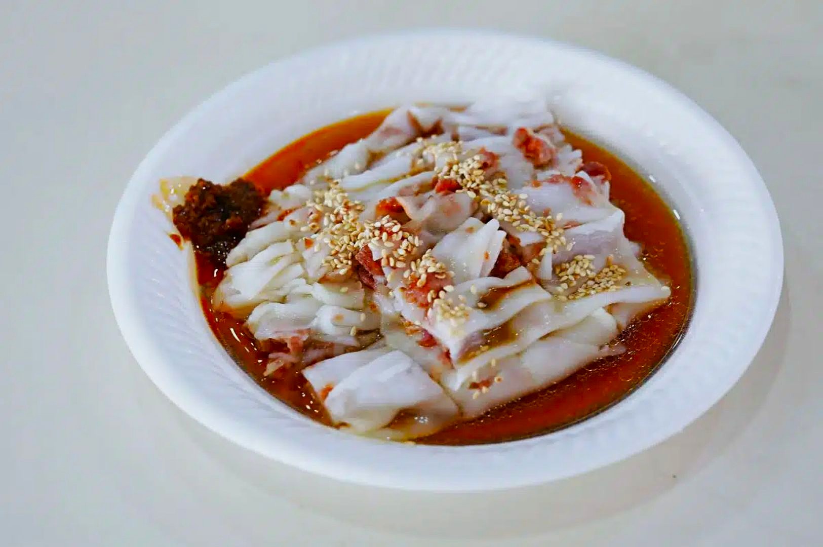Plate of chee cheong fun rice noodles topped with sweet soy sauce, bits of dried shrimp, sesame seeds, and a side of chili paste, on a white plate.
