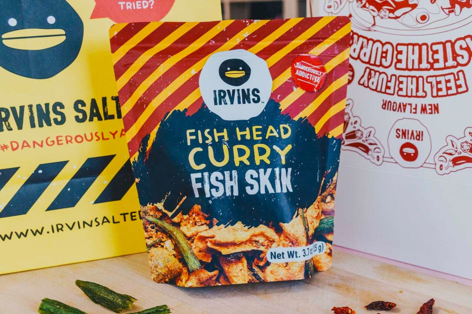 Close-up of Irvins Fish Head Curry Fish Skin packaging on a wooden surface, featuring a bold blue splash design and scattered snack pieces nearby.