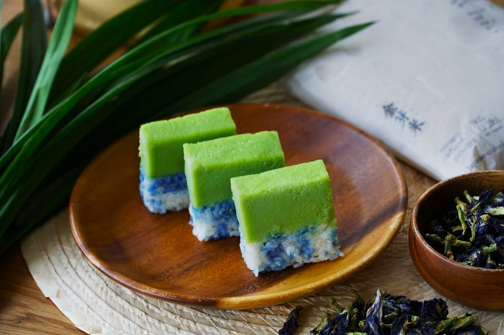 Three squares of vibrant green and blue layered cake sit on a wooden plate. Beside them are fresh and dried pandan leaves, creating a natural, fresh ambiance.