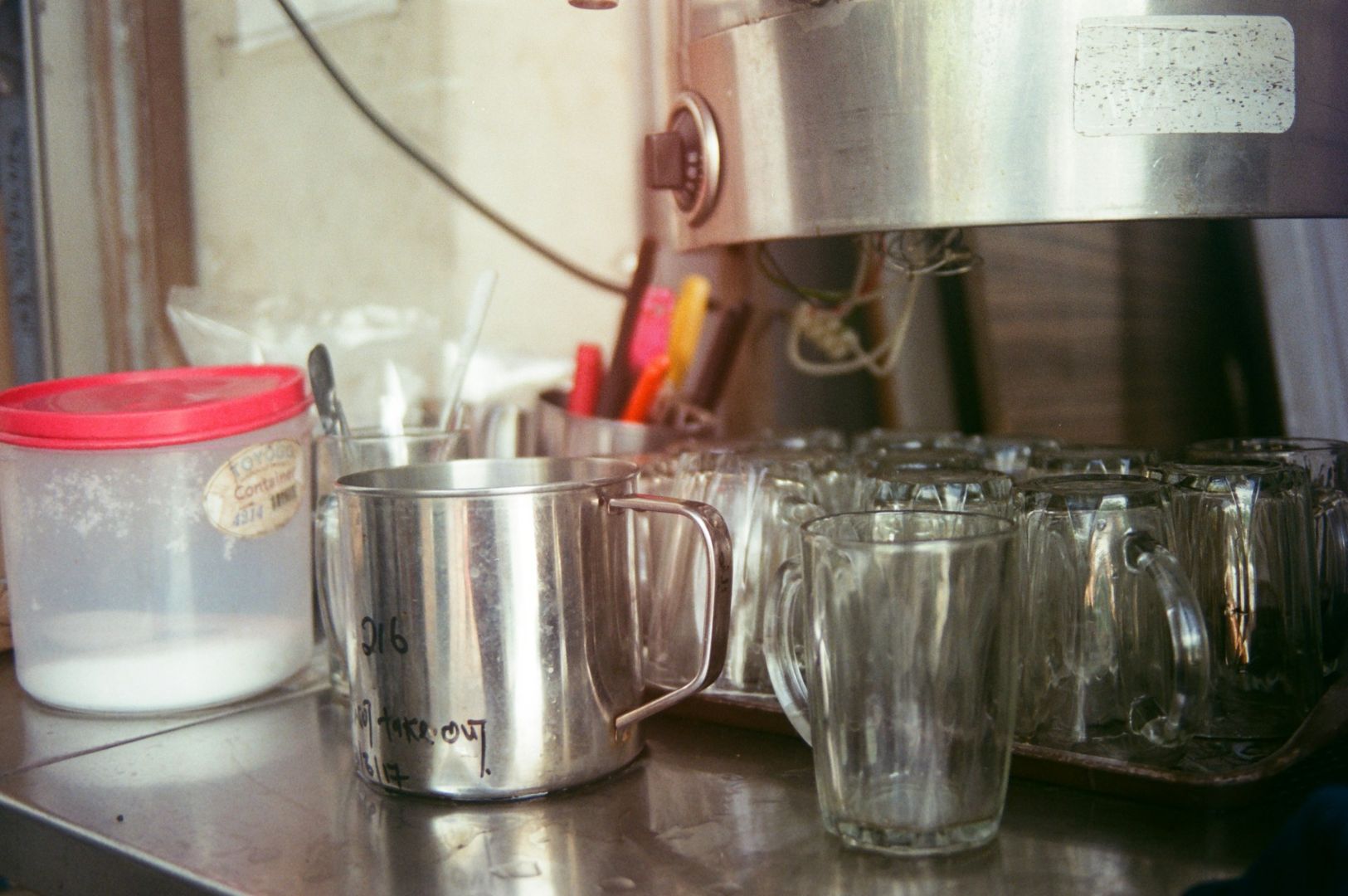 Close-up of a cluttered cafe counter with a metal frothing pitcher, glass mugs, a red-lidded container, and a coffee machine, conveying a busy atmosphere.