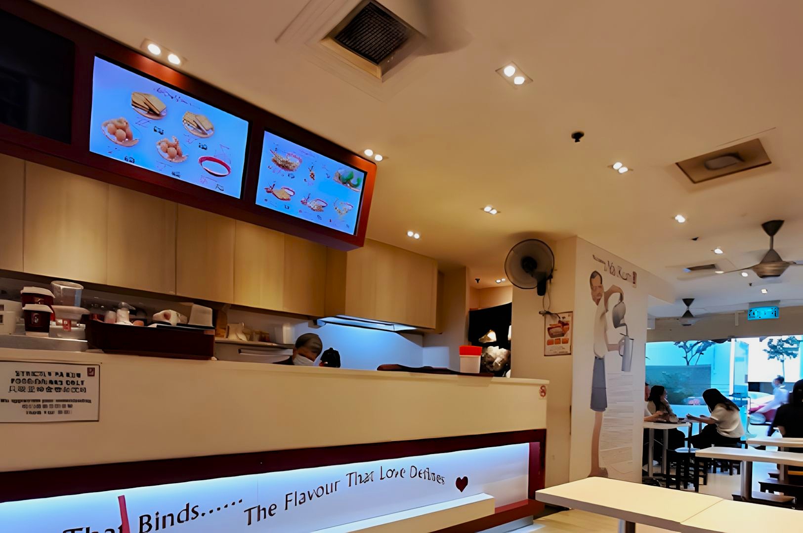 Brightly lit fast-food restaurant interior with menu screens displaying food items. Customers seated at tables. A welcoming and lively ambiance.