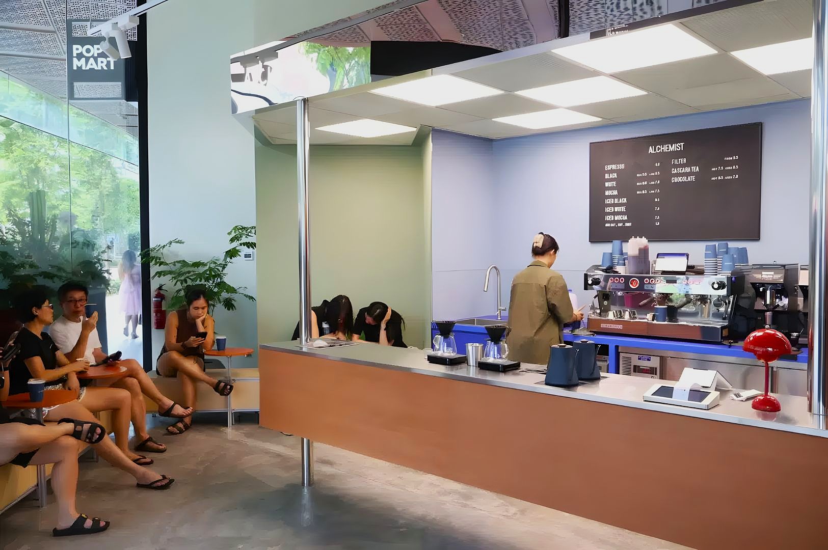 Modern coffee shop interior with a sleek design. A barista prepares drinks behind the counter, while patrons sit on the left, creating a lively atmosphere.