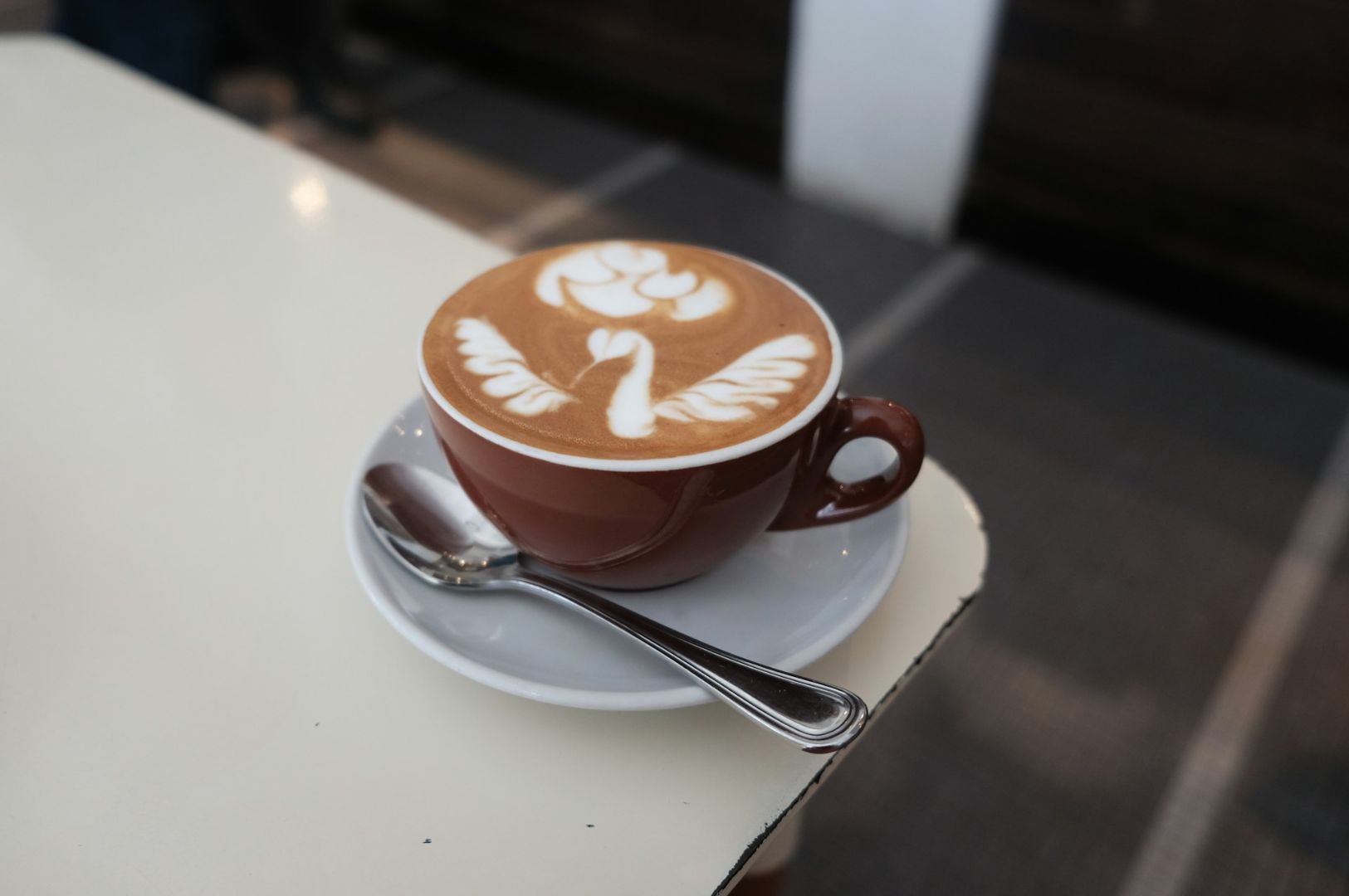 Brown cup on a white saucer with spoon, containing latte art of a swan. The cup is on a white table, creating a cozy and artistic ambiance.