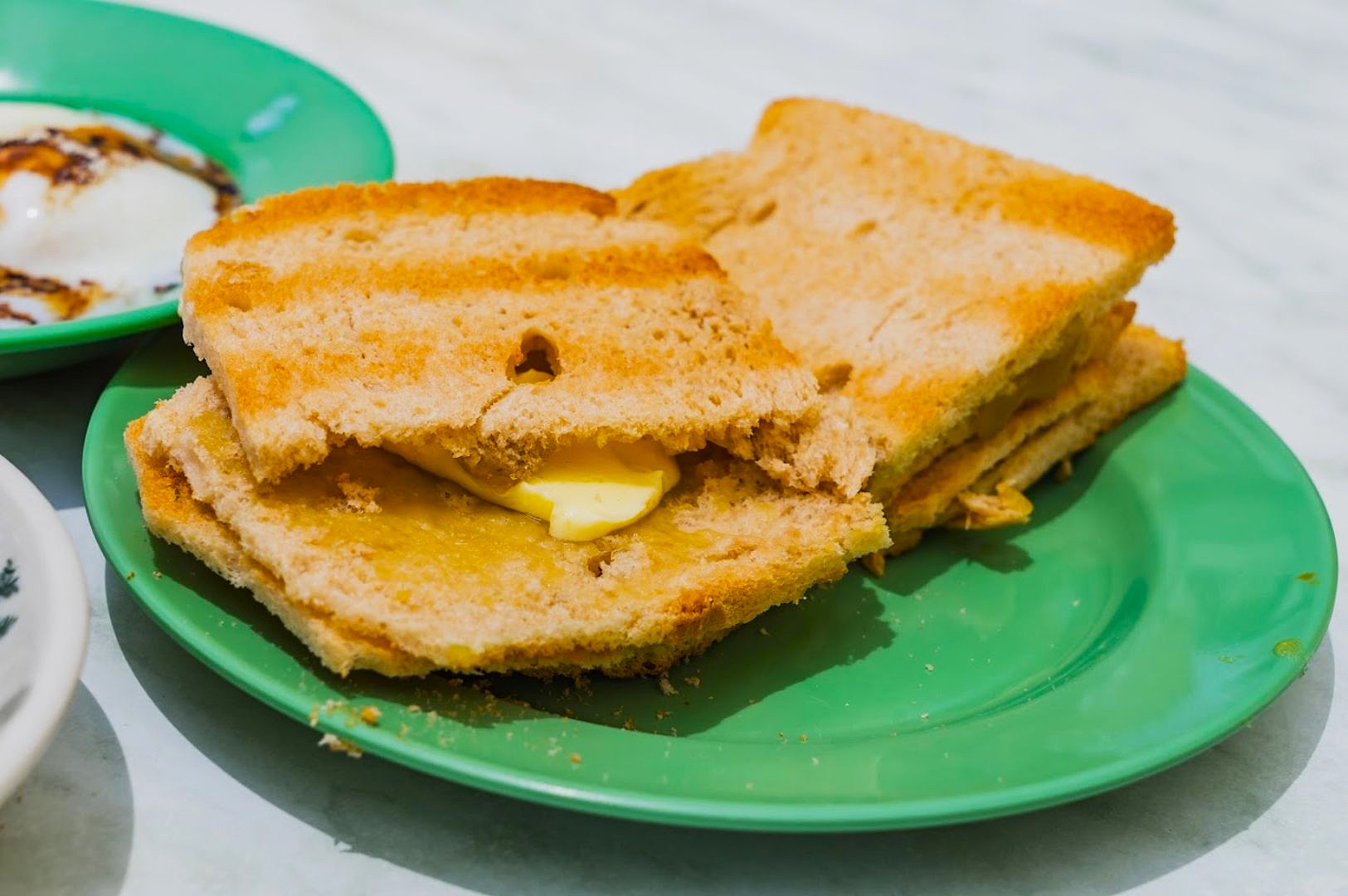 Toasted bread with soft butter on a green plate, served on a marble table. A dish with a soft-boiled egg is partially visible, evoking a classic breakfast vibe.