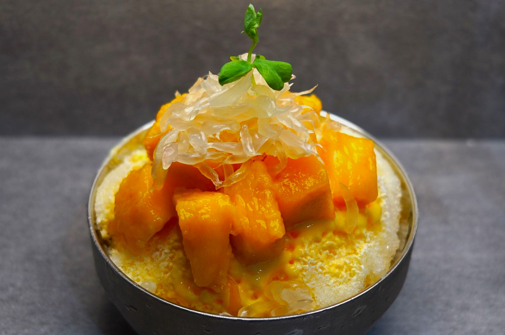 A bowl of shaved ice topped with juicy mango chunks, translucent pomelo pieces, and a green garnish, creating a fresh, vibrant dessert.