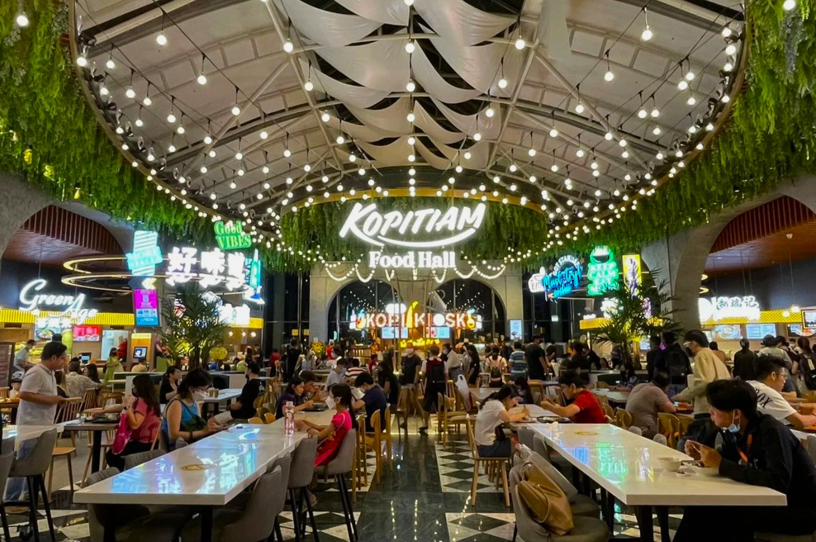 Bustling food hall with people dining at tables under a ceiling of hanging greenery and string lights. Bright neon signs and vibrant atmosphere.
