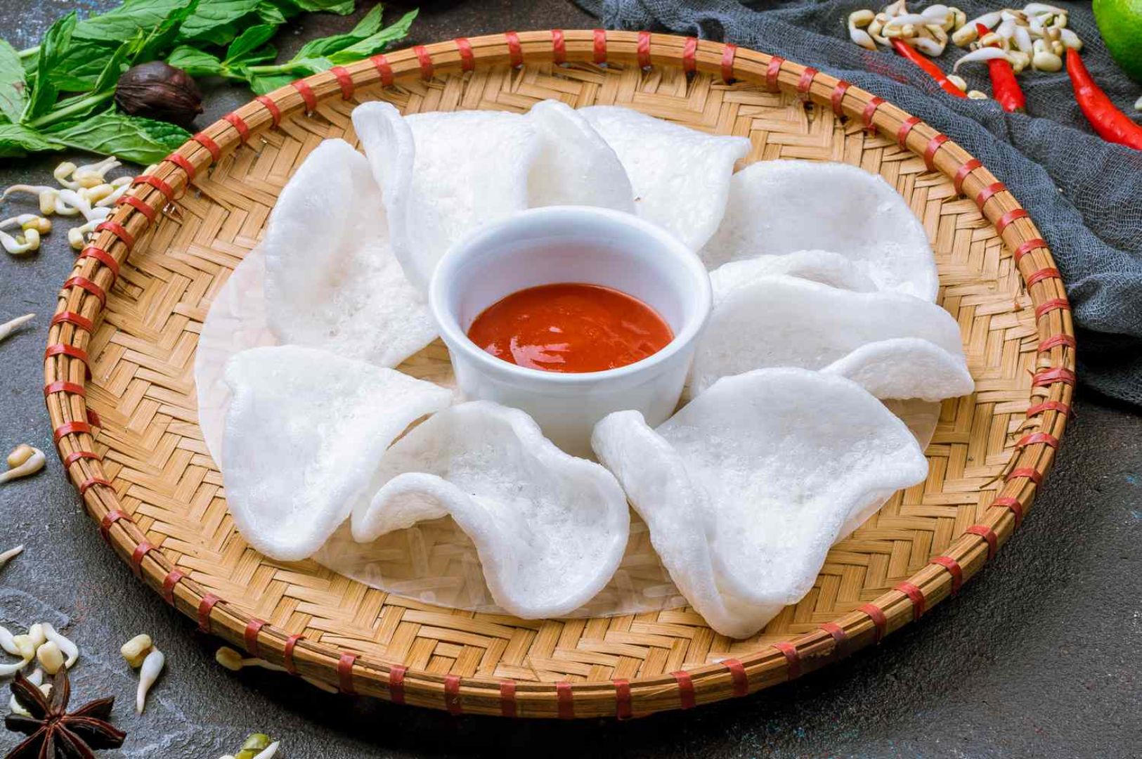 A woven bamboo tray displays white prawn crackers neatly arranged around a small bowl of red sauce. Fresh herbs and spices surround the tray, creating an inviting, appetizing tone.