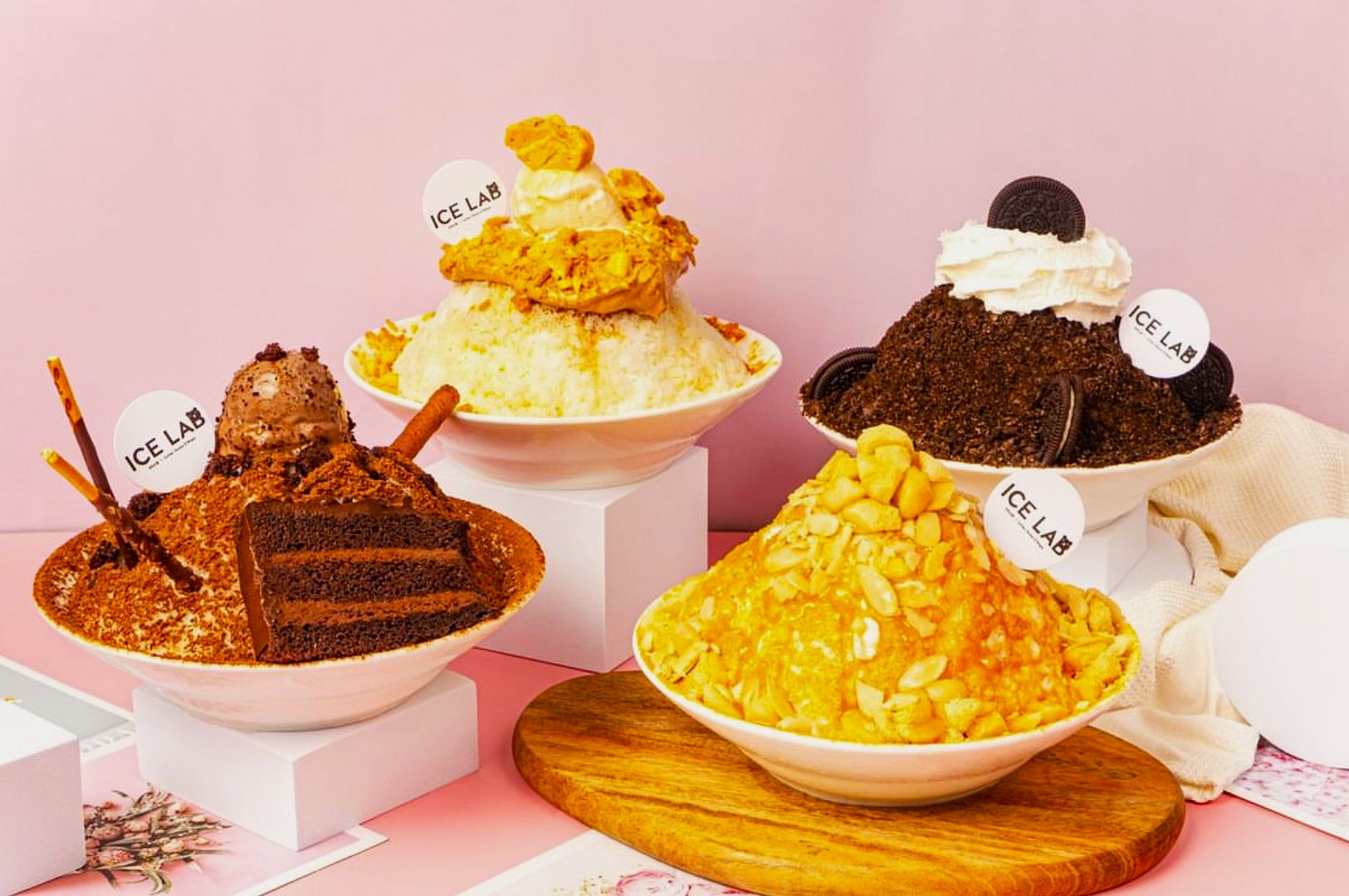 Three bowls of shaved ice desserts on a pink background. Flavors include chocolate with cake, pumpkin with custard, and cookies with whipped cream.