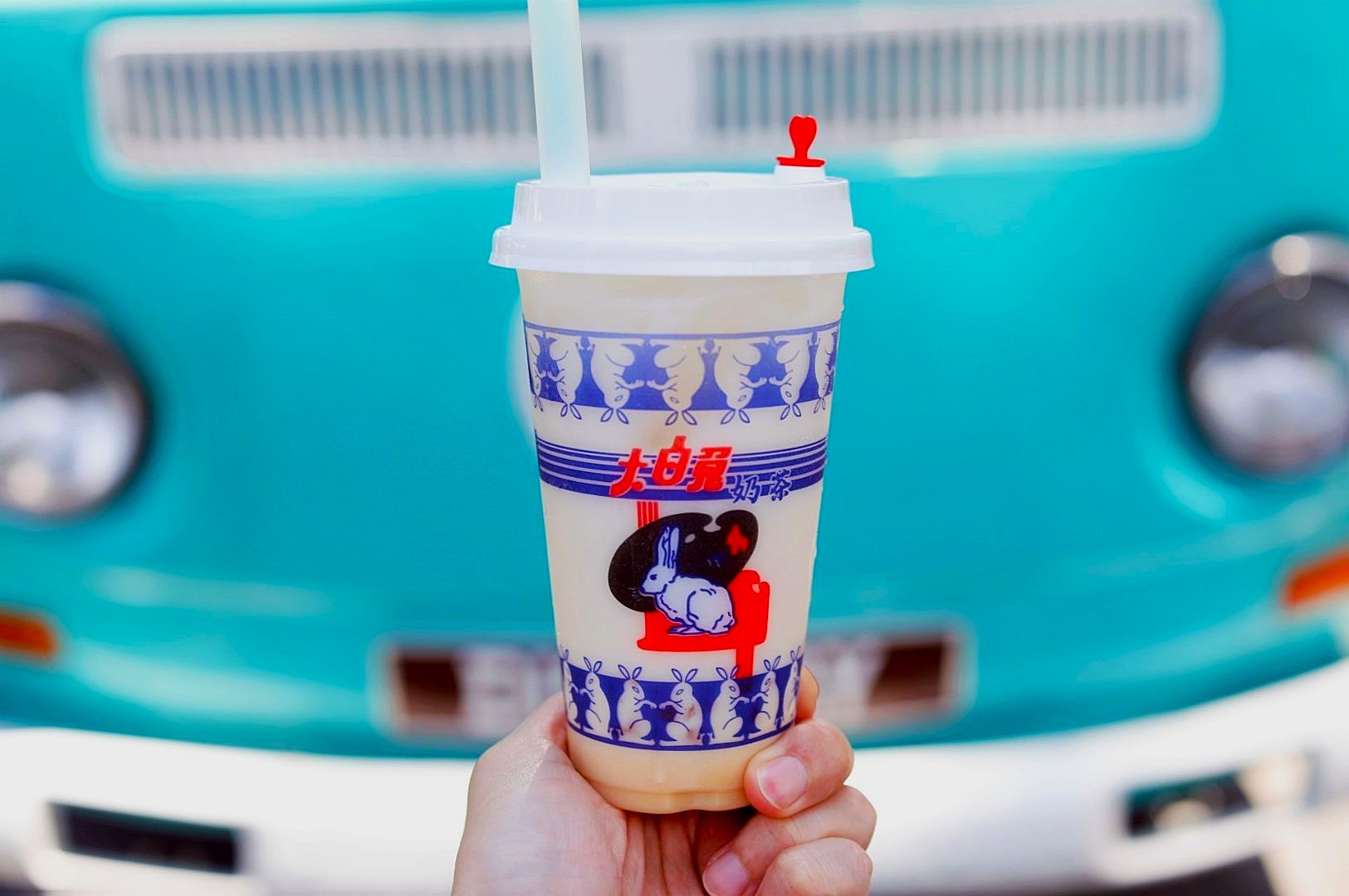 A hand holds a bubble tea cup with rabbit designs, featuring a white lid and red heart stopper. A turquoise vintage van is blurred in the background.