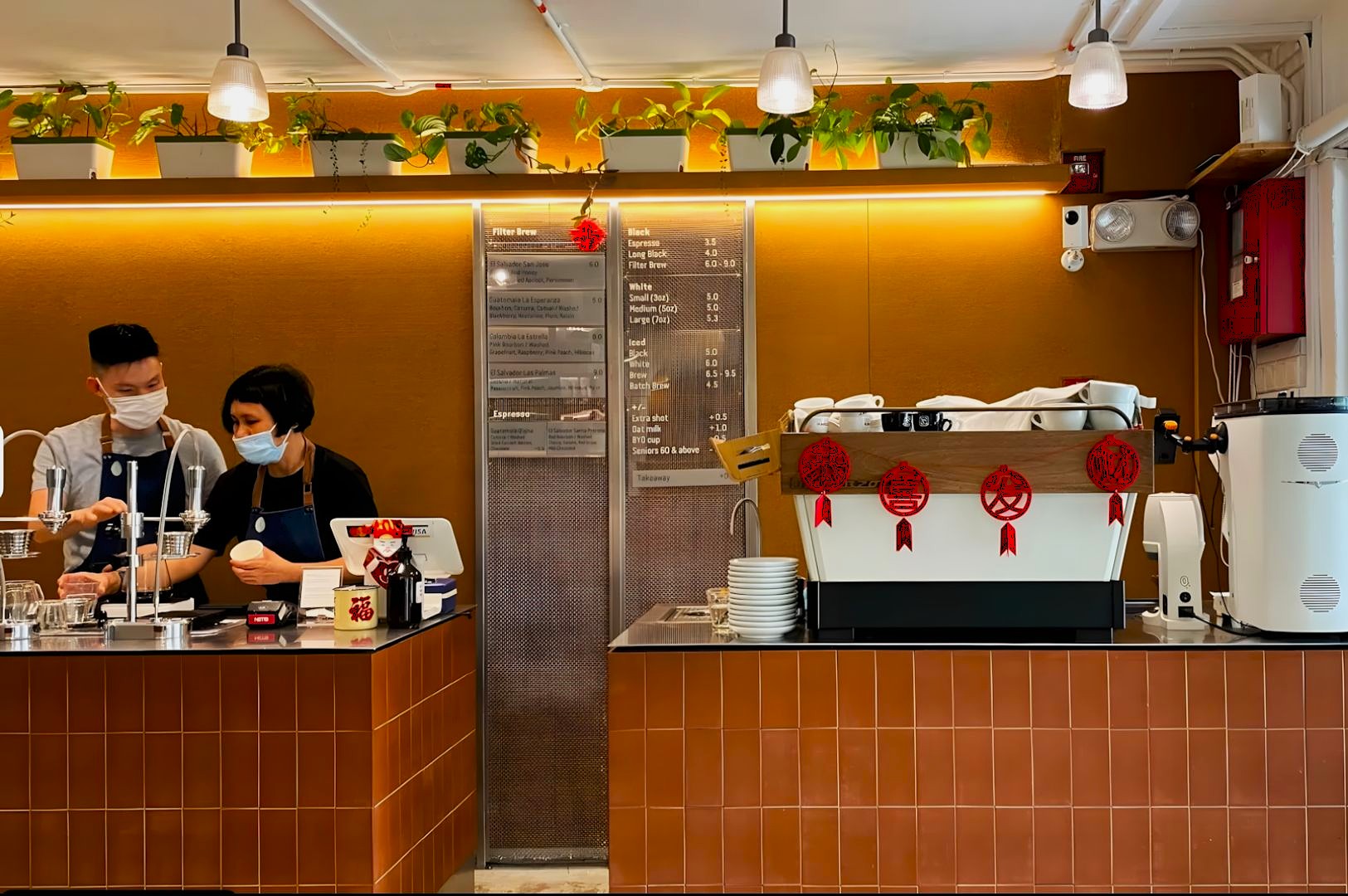 Two baristas in masks work behind a brown-tiled counter in a cozy cafe with plants and a menu board. The scene is warm and inviting.
