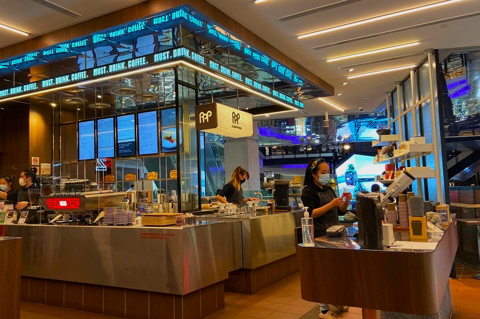 Modern coffee shop with sleek design, featuring a counter with two masked baristas. Digital menus and warm lighting create a welcoming atmosphere.