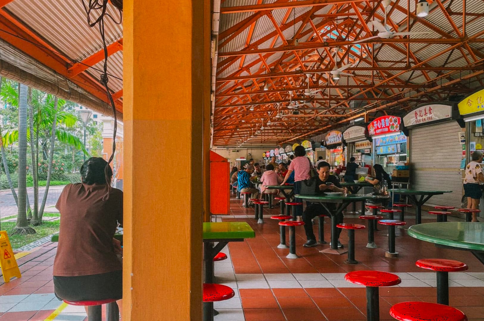 Open-air food court with red stools and green tables. People sit and eat under a metal roof. Bright signs above closed and open food stalls convey a lively atmosphere.