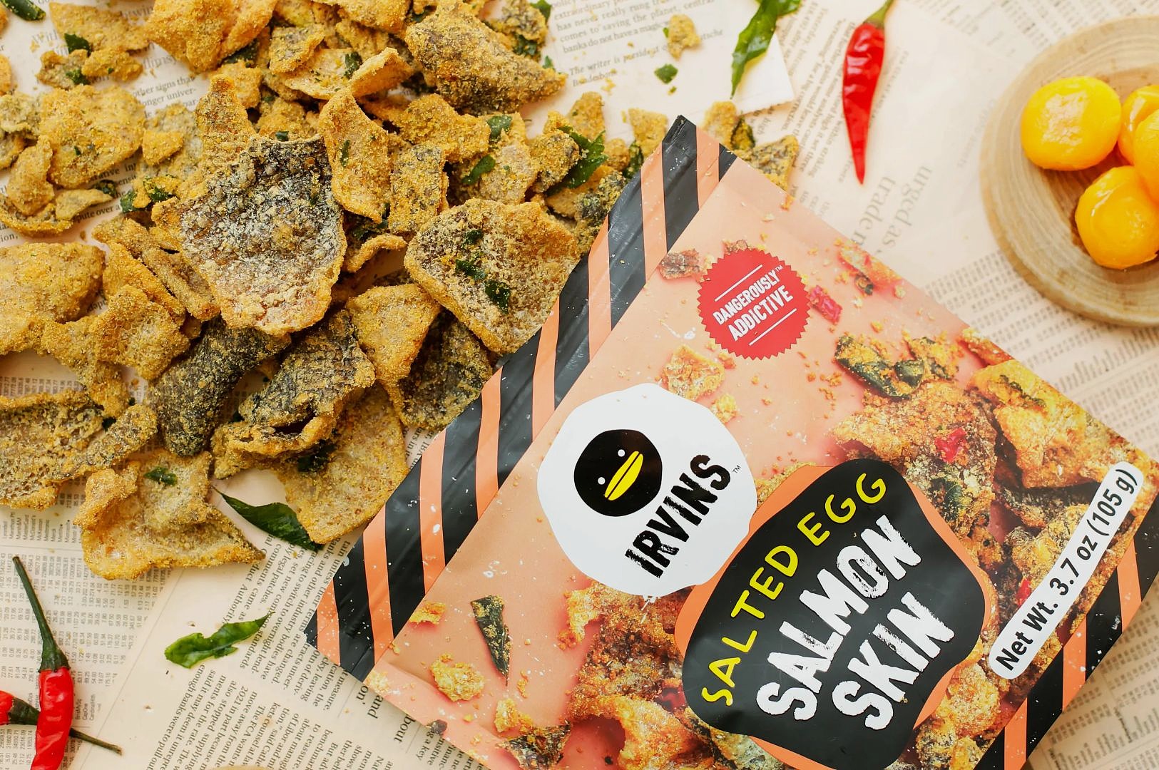 Close-up of seasoned fried fish skin crackers on paper, showing texture and herbs, alongside a pink Salted Egg Salmon Skin package.