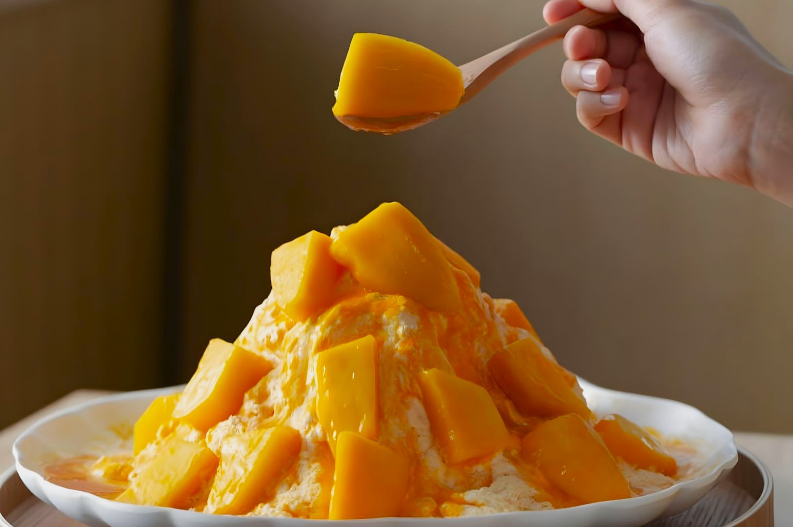 A hand holds a spoonful of mango chunks above a snow ice dessert topped with vibrant yellow mango pieces, evoking a refreshing, sweet scene.