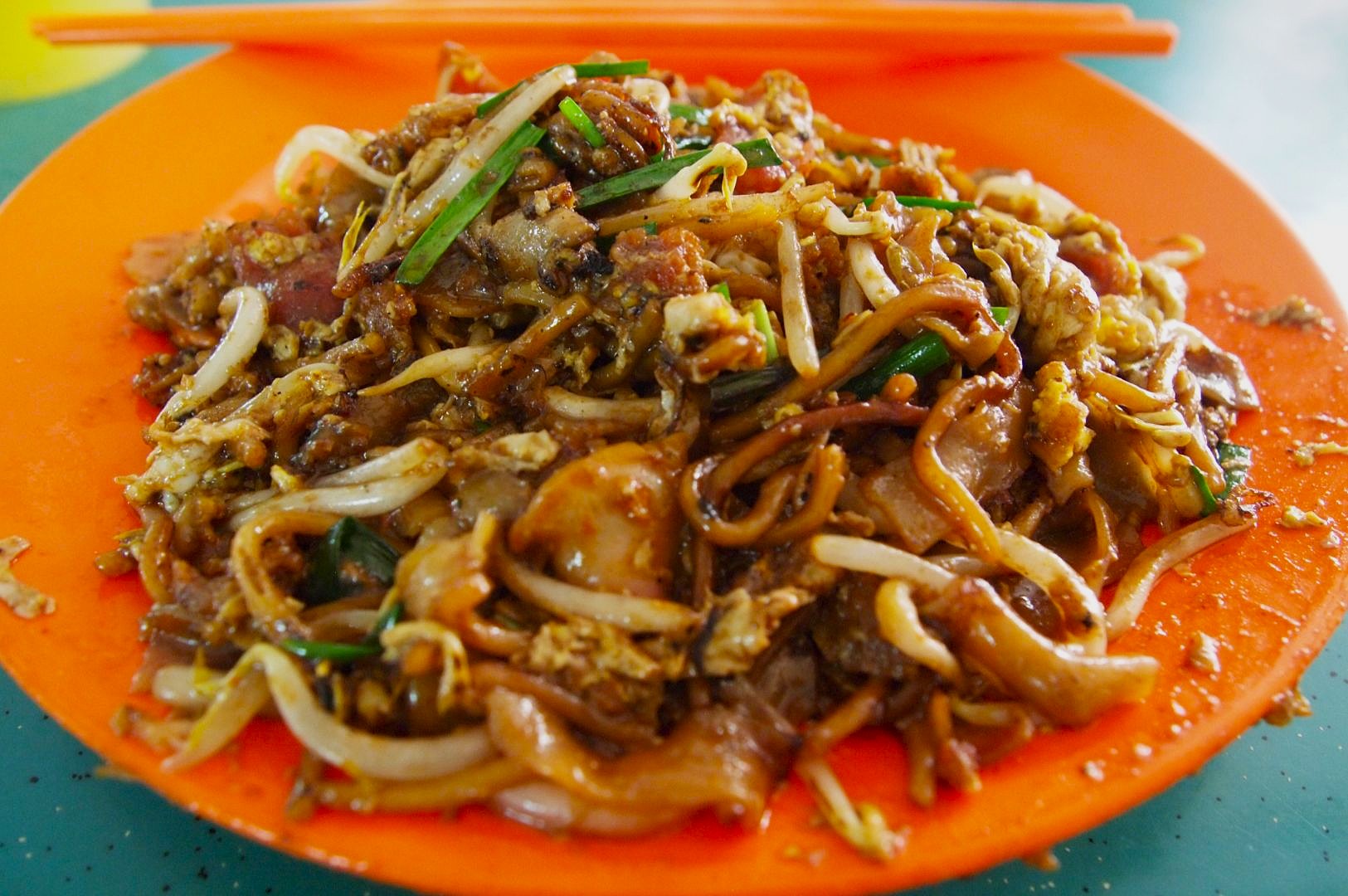 A plate of char kway teow is served on an orange dish, showcasing stir-fried noodles with bean sprouts, chives, and scrambled eggs. Bright orange chopsticks sit on top.