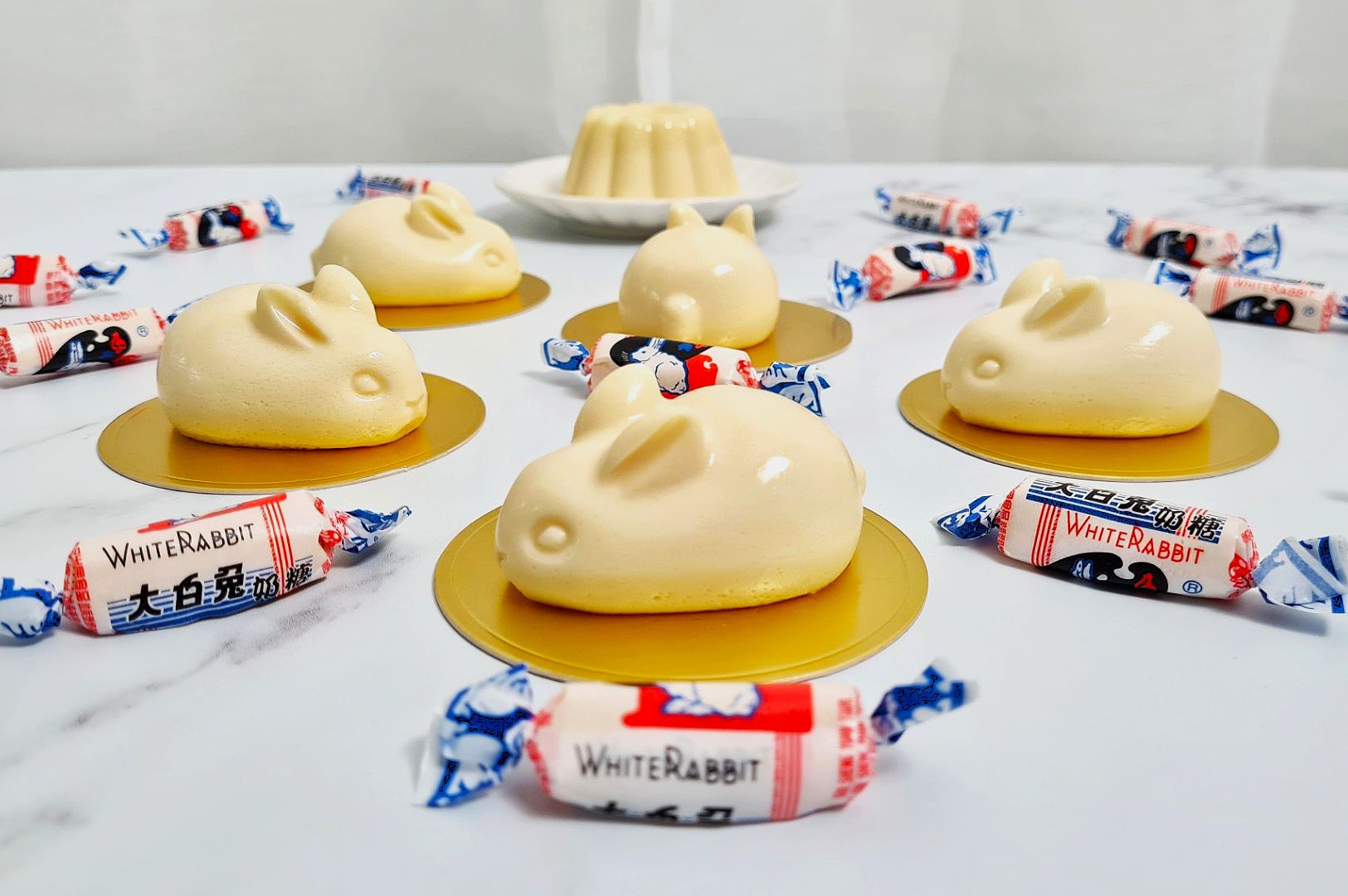 Creamy bunny-shaped desserts are displayed on gold plates, surrounded by wrapped White Rabbit candies. A flan sits in the background, creating a playful and sweet ambiance.