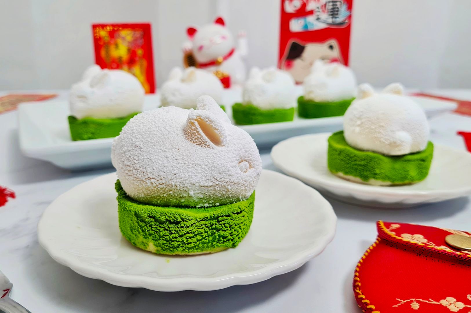 White rabbit-shaped mousse cakes on green bases are displayed on plates. A waving cat figure and festive red envelopes add a joyful, celebratory tone.