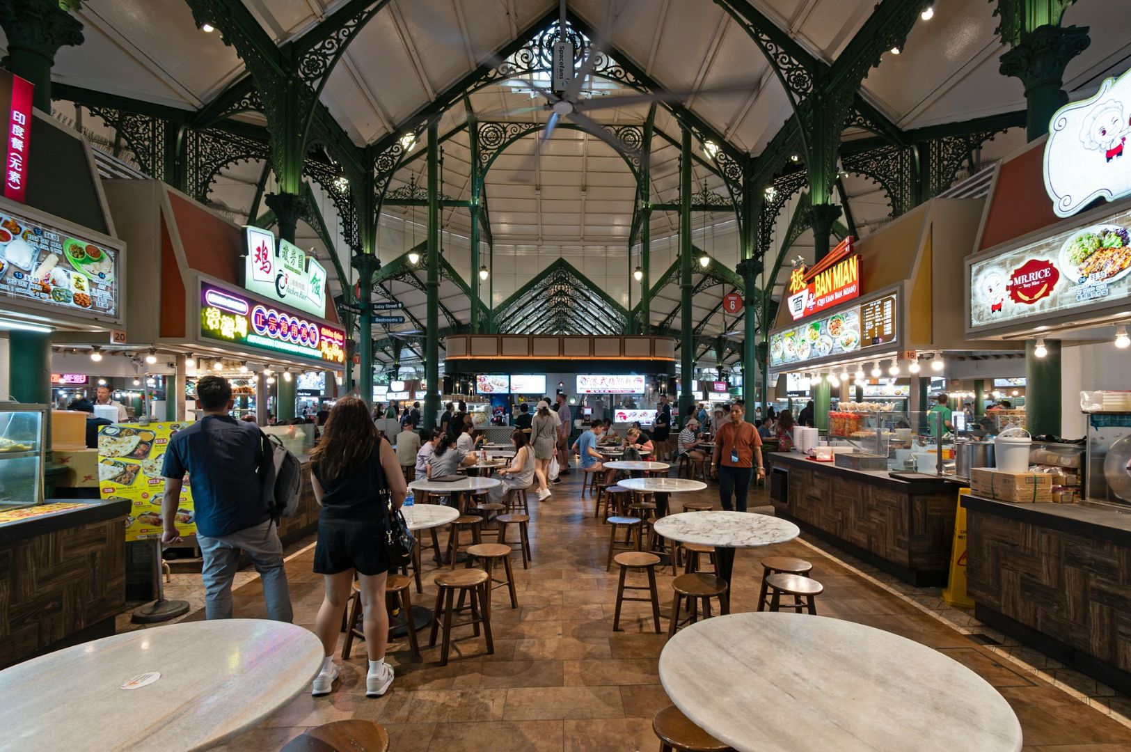 Spacious food court with ornate green ironwork. People walk past diverse food stalls beneath a high ceiling. Round tables and stools fill the center area. Vibrant, lively atmosphere.