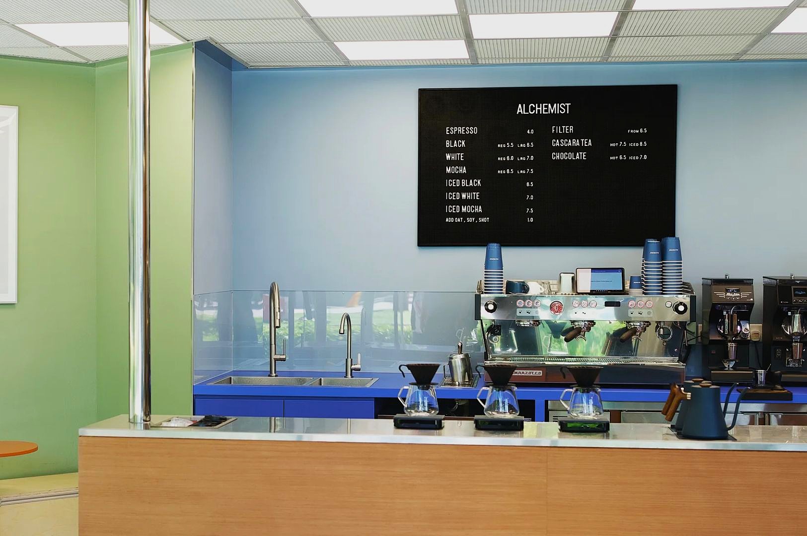 Modern coffee shop interior with a clean blue and green color scheme. A menu board lists coffee options above a sleek espresso machine and brewing station.