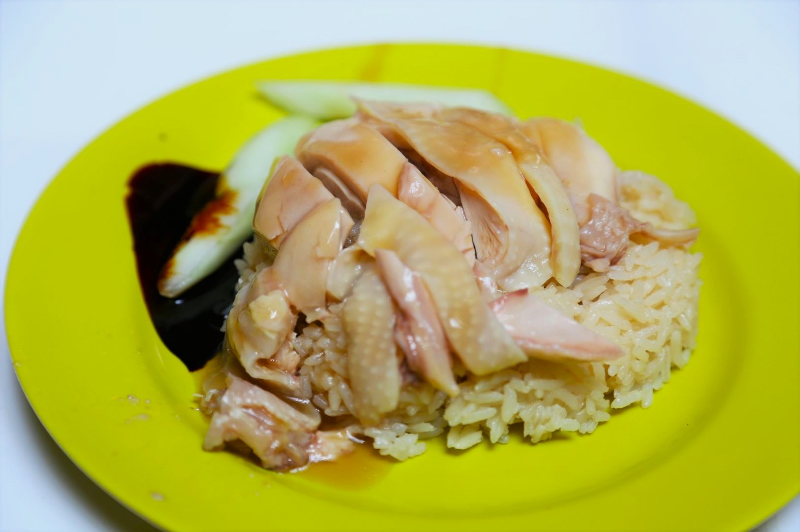 Sliced poached chicken on rice served on a yellow plate with cucumber slices and dark soy sauce. The dish looks appetizing and neatly arranged.