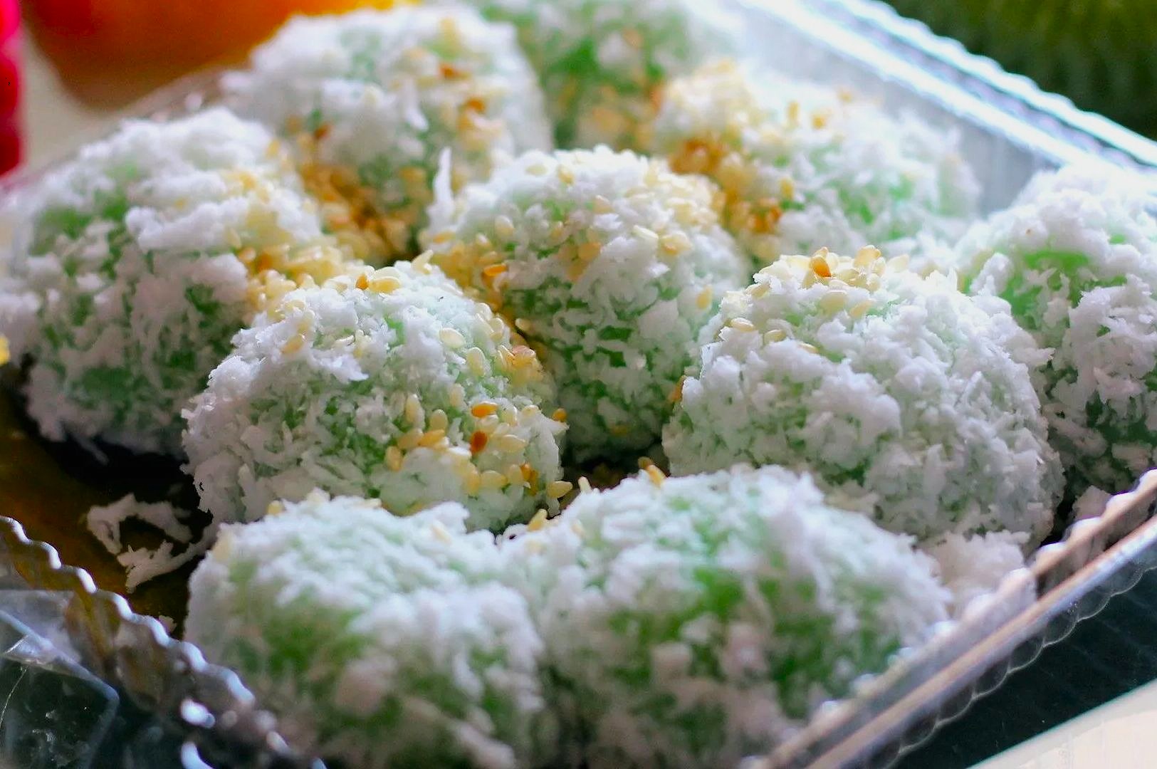 Close-up of eight green glutinous rice balls sprinkled with sesame seeds and coated in shredded coconut, arranged in a clear plastic tray. The tone is fresh and inviting.
