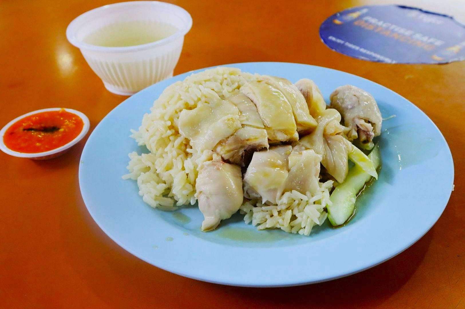 Plate of Hainanese chicken rice with sliced chicken and cucumber on a blue plate, served with chili sauce and a bowl of soup on a brown table.